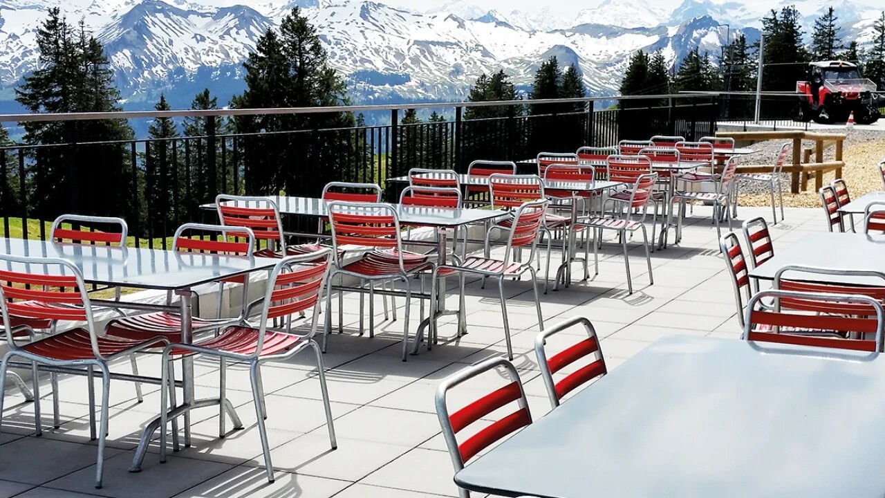 Eine Terrasse vor einem Panorama mit Tannen in der Nähe und schneebedeckten Bergen in der Ferne. Die roten Outdoor-Stühlen haben ein verchromtes Gestell sind Manufakt und die grauen Tische sind von Schaffner.